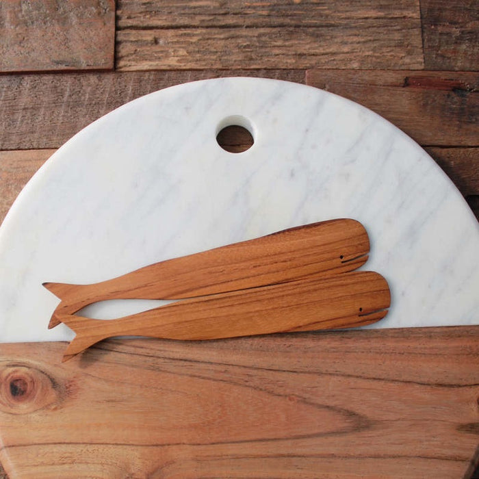 The BE HOME - TEAK WHALE SPREADERS by BE HOME, INC rest on a round white marble cutting board with a hole at the top, displayed on a wooden surface.