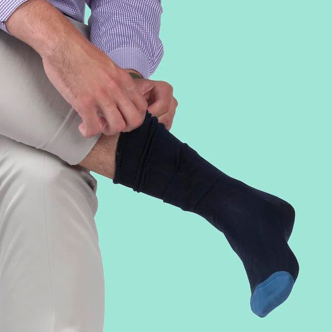 A person in light pants and a purple checkered shirt adjusts BIRD DOG BAY - PEDIGREE OVER THE CALF SOCKS IN BLUE while sitting against a light turquoise background.