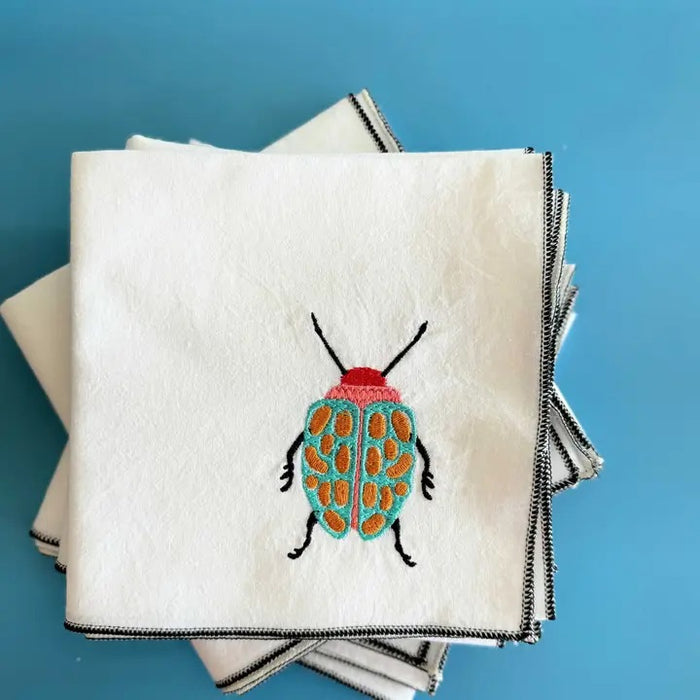 The DOT AND ARMY - EMBROIDERED BEETLE TOWEL by DOT AND ARMY is a neatly folded white cotton kitchen towel with black trim, embroidered with a colorful beetle featuring a red head and turquoise body with orange spots.