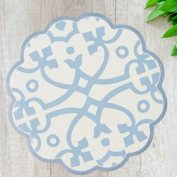 The ETUHOME Round Scalloped Placemat in Denim, featuring a blue and white geometric floral pattern, is displayed on a light wooden surface with green plant leaves visible in the upper right corner.