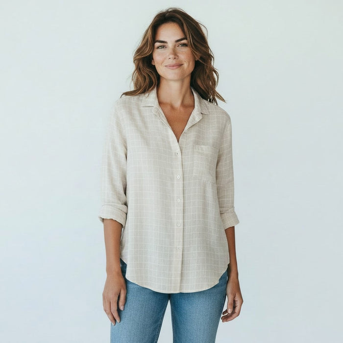 A woman with brown hair, smiling on a sunlit cobblestone street with blurred outdoor café tables in the background, wears the FRANK & EILEEN EILEEN Relaxed Button Up in Italian Cotton Tan with Textured Grid paired with jeans.