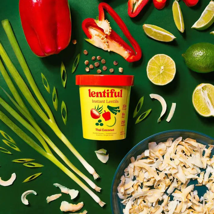 A yellow container of LENTIFUL - THAI COCONUT CURRY LENTILS is surrounded by fresh red bell pepper, lime halves, green onions, coconut flakes, and sliced chili on a green background, highlighting a spicy meal from LENTIFUL.