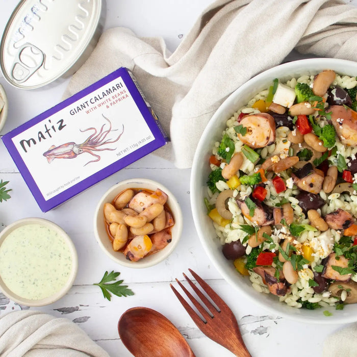 A bowl of rice salad with vegetables and wild-caught calamari sits beside a box labeled “Matiz España - Giant Calamari with White Beans and Paprika,” a small bowl of this gourmet seafood from Matiz España, a wooden fork and spoon, and a dish of green sauce infused with Spanish paprika on a white table.