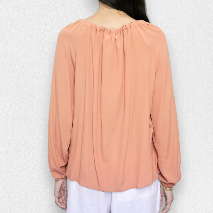 A woman in the MEIMEIJ Tie Neck Blouse—loose, long-sleeved, peach-colored washable silk with a neckline tie—paired with white pants, stands against a light textured background. Her face is partially out of frame.