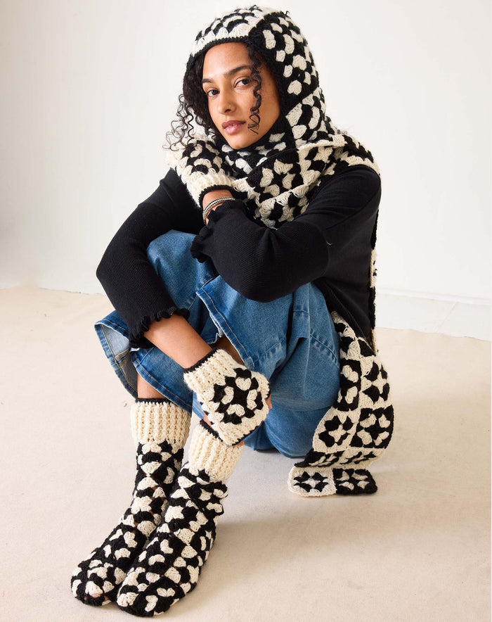 A pair of MERSEA - LA PAZ Crochet Fingerless Gloves in cream, featuring hand-crocheted black geometric patterns and black trim, displayed on a white background.