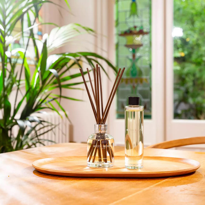 A clear glass bottle labeled "MOLTON BROWN - ORANGE AND BERGAMOT DIFFUSER" contains yellow liquid and several reed sticks, gently dispersing a citrus scent, set against a plain white background.