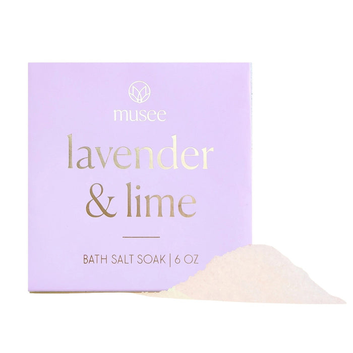 A lavender-colored box labeled “MUSEE - LAVENDER & LIME MINI BATH SALT SOAK” sits beside a small mound of white bath salts, infused with calming lavender essential oil for a soothing soak.