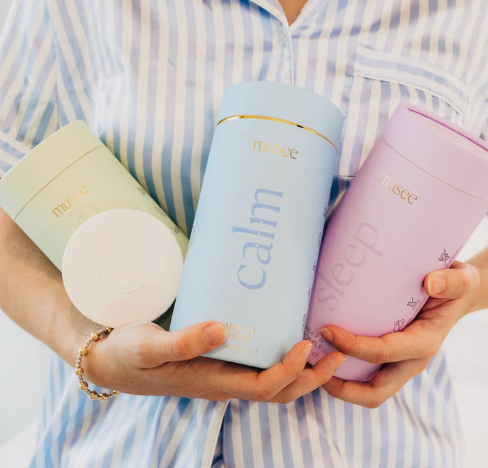 A hand wearing a gold bracelet places a MUSEE - SLEEP BATH TABLET into a tub of water, releasing gentle bubbles that rise to the surface. Green leaves in the corner highlight the natural ingredients in this bath product from MUSEE.