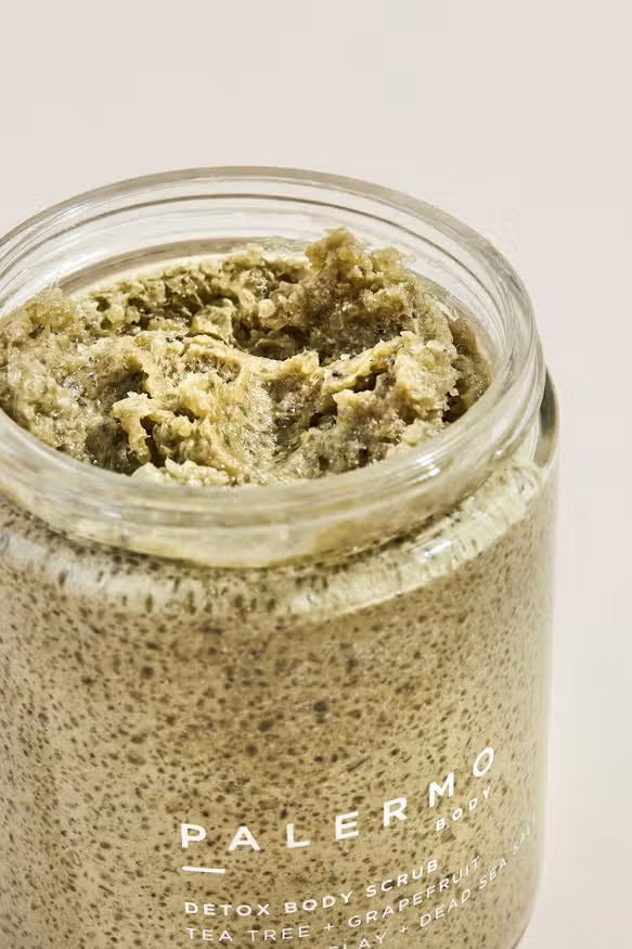A clear glass jar with a black lid holds PALERMO BODY’s TEA TREE AND GRAPEFRUIT BODY SCRUB, featuring sea clay and Dead Sea salt. The label lists 8.5 oz (240g) of detoxifying exfoliant inside.