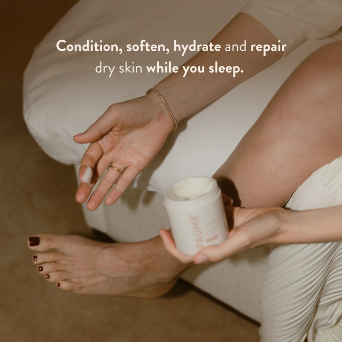 A white cylindrical container of PAUME’s OVERNIGHT HAND AND FOOT HYDRATION MASK rests on a creamy textured surface, with nourishing mask—ideal for dry, cracked skin—smeared around it.