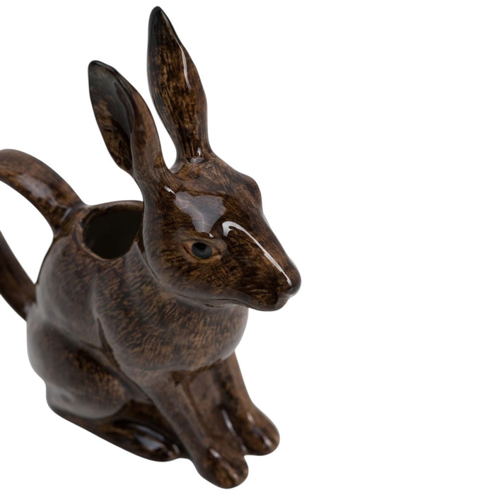 The QUAIL CERAMICS - HARE JUG, LARGE by QUAIL DESIGNS, a delightful ceramic pitcher shaped like a brown rabbit, sits on a wooden surface with white flowers artfully arranged in its open mouth. This charming decorative piece brings whimsy to the tabletop display, while green plants create a softly blurred background.