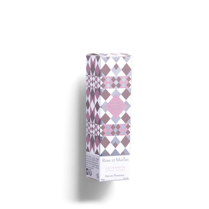A rectangular glass perfume bottle with a silver spray nozzle and square cap beside it casts a shadow on white. The label reads "ROSE ET MARIUS - A SIESTA IN A SUNLIT HOME EAU DE PARFUM 30ML," evoking the scent of Provence.