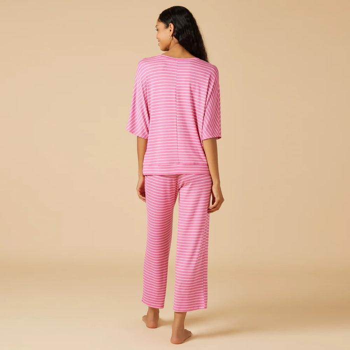 A woman stands barefoot against a beige background, wearing the SOFTIES - DREAM RELAXED V-NECK WITH CAPRI LOUNGE SET IN FUSCHIA STRIPE, featuring pink and white stripes, a loose top, and drawstring capri pants.