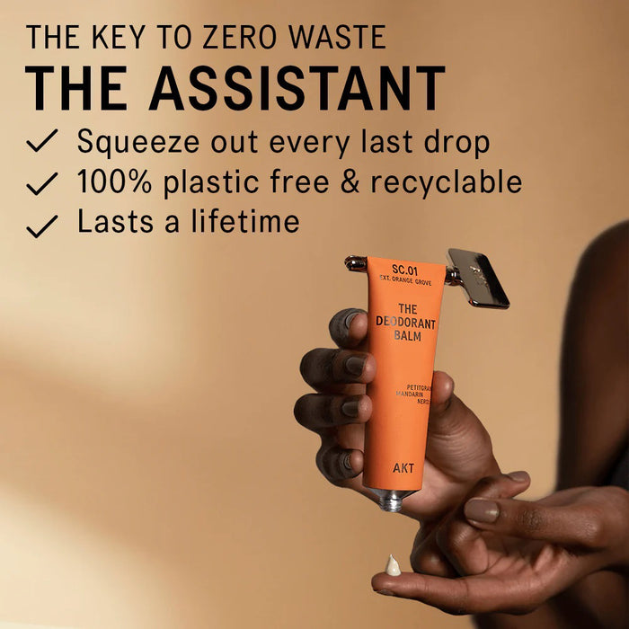 A gold THE ASSISTANT KEY TOOL by AKT LONDON and its beige, rectangular box are displayed on a smooth surface with soft sunlight and shadows, showcasing this elegant, plastic-free addition to your routine.