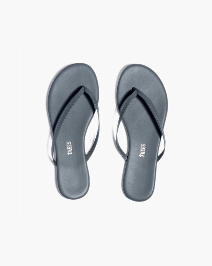 A pair of TKEES INC LILY LINER SLIPPER ASH black flip-flops with thin straps, made from Brazilian leather, are placed side by side on a plain white background with the "TKEES" logo visible on the cushioned insole.
