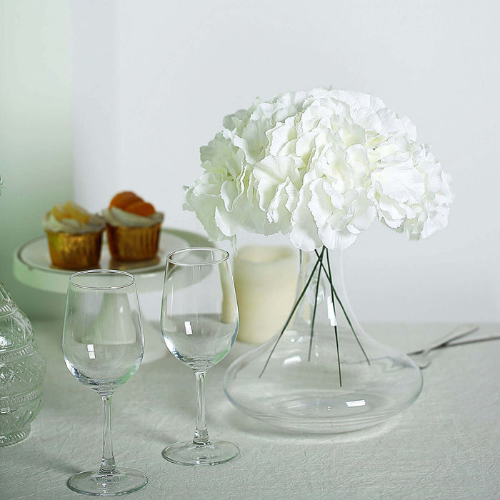 White hydrangea bouquet in a clear vase with two wine glasses on a table.