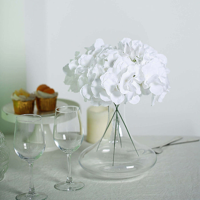 White hydrangea bouquet in a glass vase with wine glasses on a table.