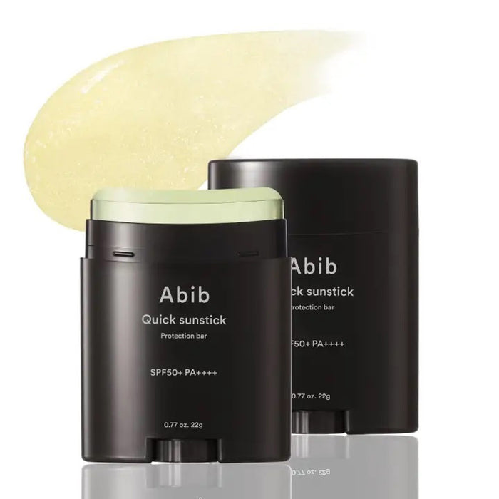Two black ASIAN BEAUTY WHOLESALE QUICK SUNSTICK PROTECTION BAR 22G are shown: one open, revealing the pale yellow stick, the other closed. A swatch of the portable sunscreen is smeared above them on a white background.