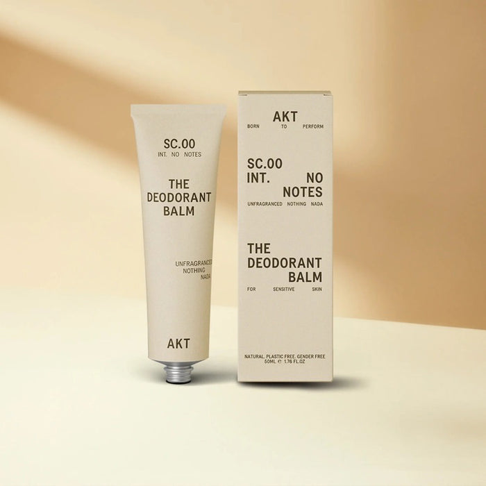 A beige tube of AKT LONDON’s AKT DEODORANT BALM FRAGRANCE FREE for sensitive skin stands upright beside its matching box on a neutral background; both display minimalist black text and design.