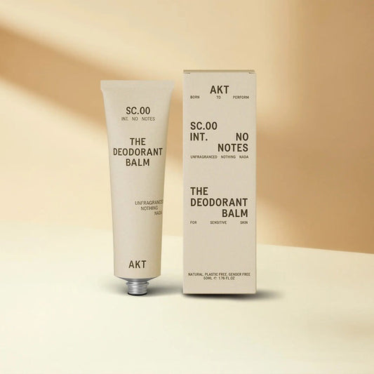 A beige tube of AKT LONDON’s AKT DEODORANT BALM FRAGRANCE FREE for sensitive skin stands upright beside its matching box on a neutral background; both display minimalist black text and design.