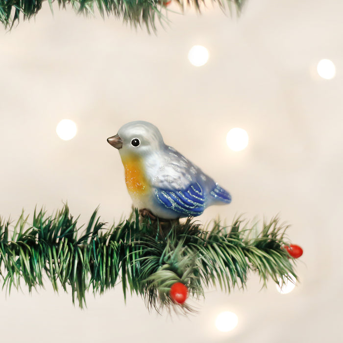 A blue, silver, and yellow bird ornament sits on a green pine branch with red berries, against a softly blurred background with glowing white lights.