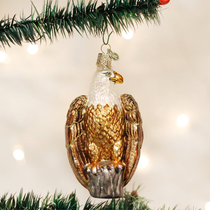 A glass Christmas tree ornament shaped like a bald eagle with gold and brown feathers, perched on a branch, hangs from a green pine branch against a blurred, light background.