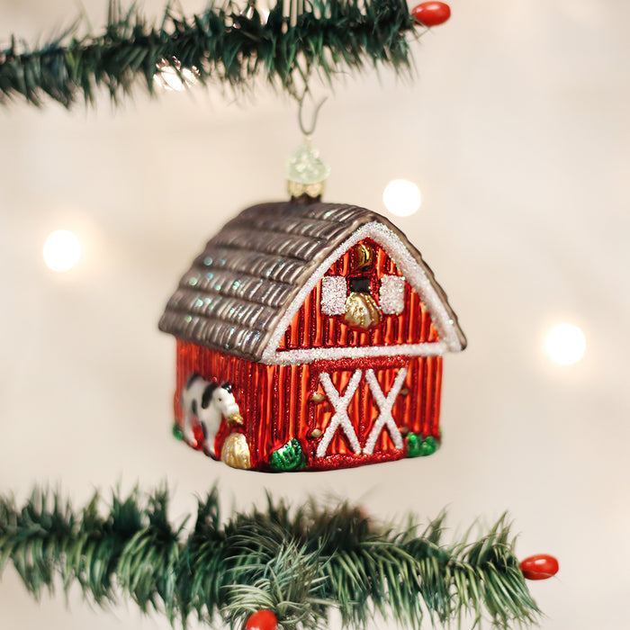 A red barn ornament with white details and a small horse stands on a decorated Christmas tree branch, surrounded by festive lights.