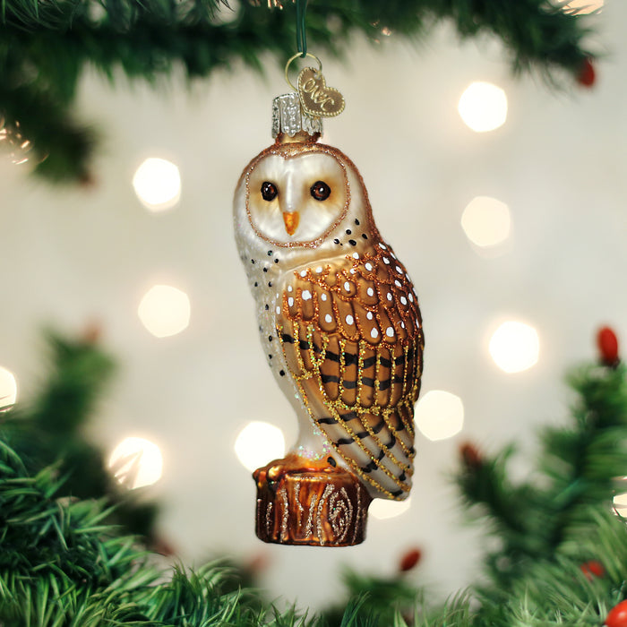 A glass owl ornament with brown and white details hangs from a Christmas tree branch, surrounded by blurred white holiday lights and green pine needles.
