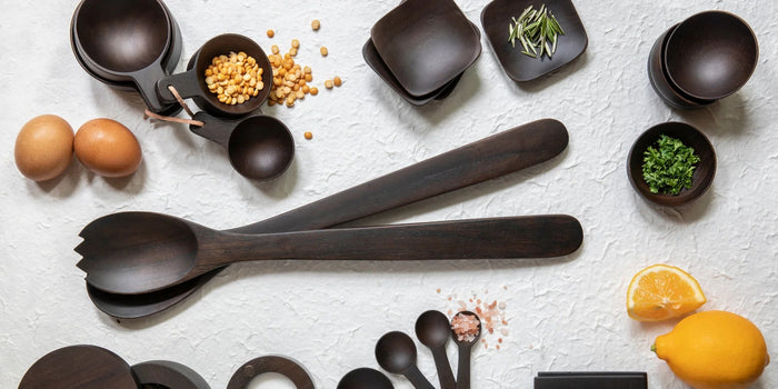 The BE HOME - Ebony Teak Streamlined Serving Set by BE HOME, INC features two hand-carved dark wooden utensils—a spoon and a jagged-edge spork—each with long handles, thoughtfully placed side by side on a white background. These elegant ebony teak servers add a modern touch to any dining table.