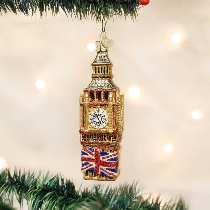 A Christmas tree ornament shaped like Big Ben hangs from a branch. The ornament is gold with a clock face and features a glittery Union Jack flag at its base. Out-of-focus lights create a festive background.