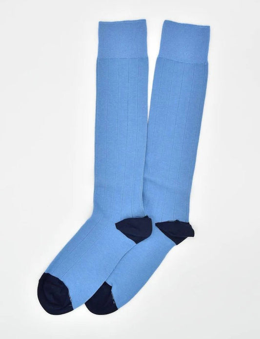 A person in light pants and a purple checkered shirt adjusts BIRD DOG BAY - PEDIGREE OVER THE CALF SOCKS IN BLUE while sitting against a light turquoise background.