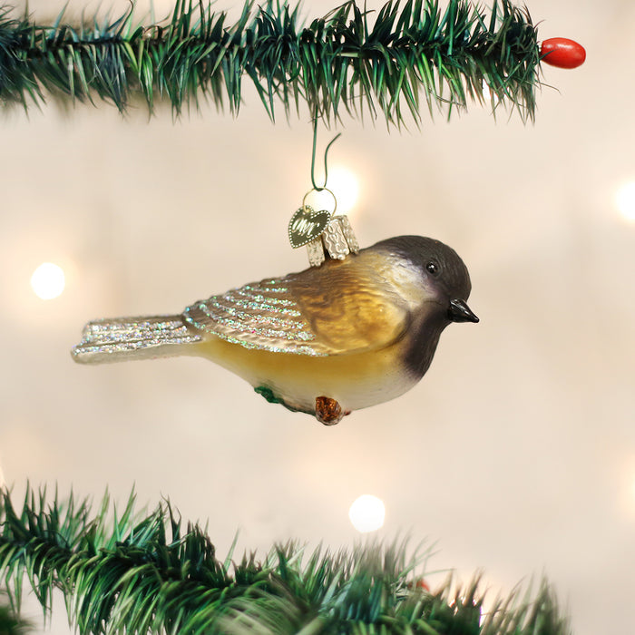 A bird-shaped ornament with brown, white, and yellow coloring hangs from a green Christmas tree branch. Soft lights glow in the blurred background.