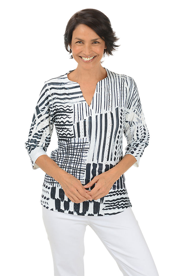 Woman wearing a 3/4 sleeve black and white patterned blouse with white pants on a white background.