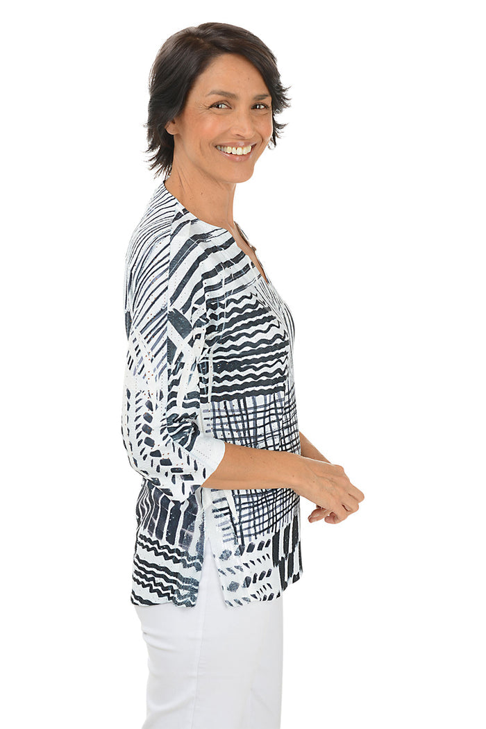 Woman wearing a 3/4 sleeve black and white patterned blouse with white pants on a white background. Side shot.
