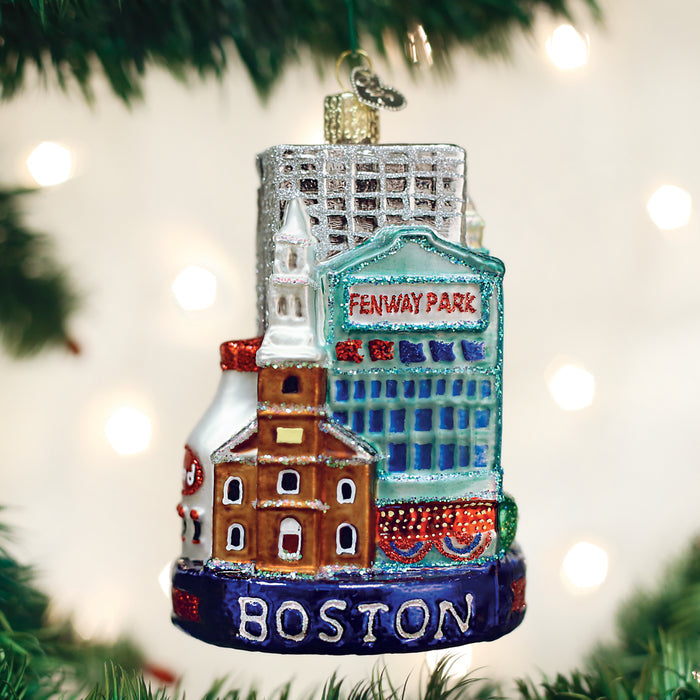 A Christmas tree ornament featuring Boston landmarks, including Fenway Park and historic buildings, with the word BOSTON at the base, hangs on a decorated tree with blurred lights in the background.