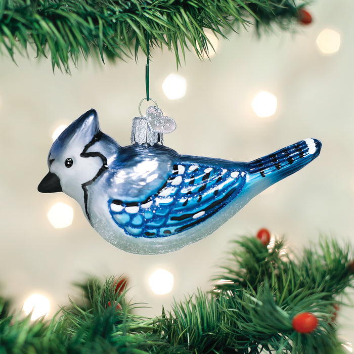 A glass ornament shaped like a blue jay bird hangs from a green Christmas tree branch with red berries, set against a softly blurred background with white lights.