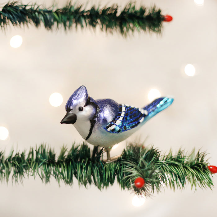 A blue jay ornament with shimmering blue and white feathers perched on a green artificial pine branch with red berries, set against a blurred background of white holiday lights.