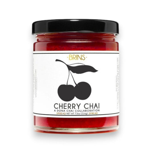 A glass jar of BRINS - CHERRY CHAI SPREAD & PRESERVE features a black lid and a label adorned with an illustration of two cherries alongside flavor details and collaboration notes. Made by BRINS with locally sourced New York sour cherries, this preserve is expertly infused with aromatic chai spices for a distinctive and flavorful experience.