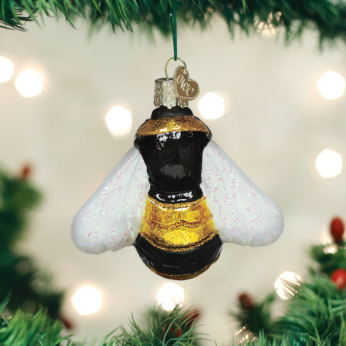 A glass ornament shaped like a bumblebee with gold, black, and white details hangs on a Christmas tree, surrounded by green branches and blurred white lights in the background.
