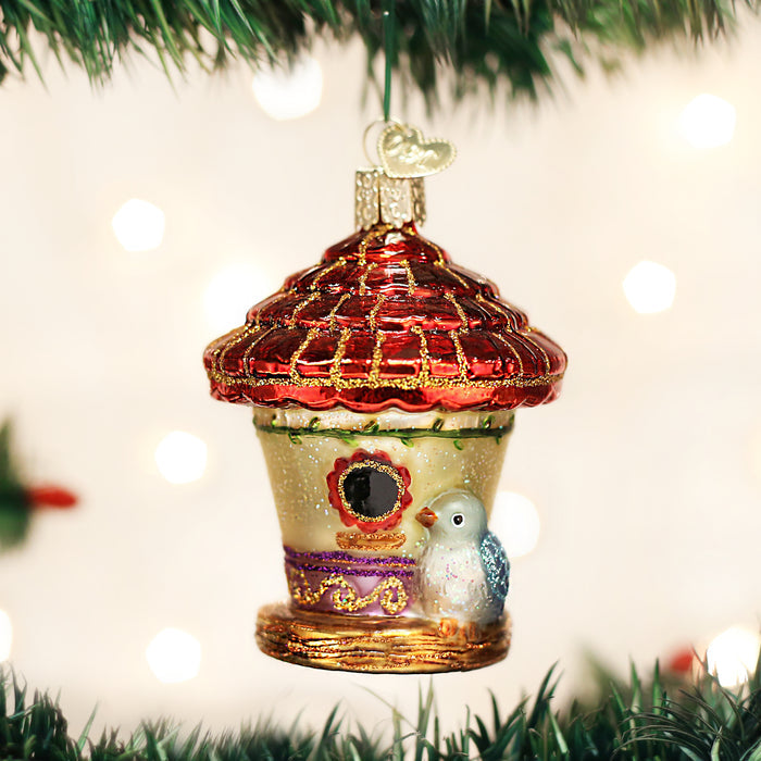 A colorful birdhouse Christmas ornament with a red roof and a small bird sits on a decorated tree branch, surrounded by blurred holiday lights in the background.