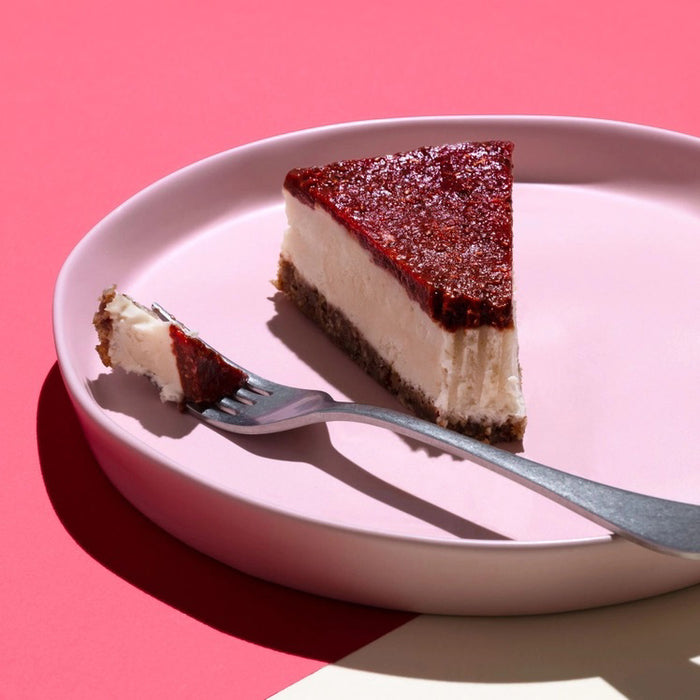 A slice of cheesecake topped with CHIA SMASH - RASPBERRY CHIA JAM by CHIA SMASH sits on a light pink plate, with a fork holding a bite nearby. The scene features a bright pink background and bold shadows.