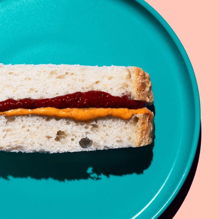 A slice of cheesecake topped with CHIA SMASH - RASPBERRY CHIA JAM by CHIA SMASH sits on a light pink plate, with a fork holding a bite nearby. The scene features a bright pink background and bold shadows.