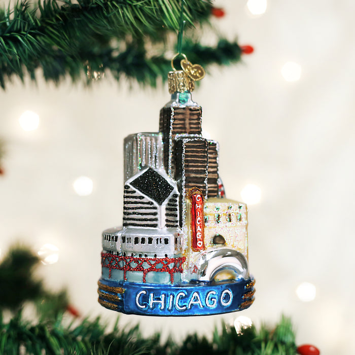 A Christmas tree ornament shaped like Chicago skyscrapers, decorated with glitter and the word CHICAGO at the base, hangs on a green branch with blurred white lights in the background.