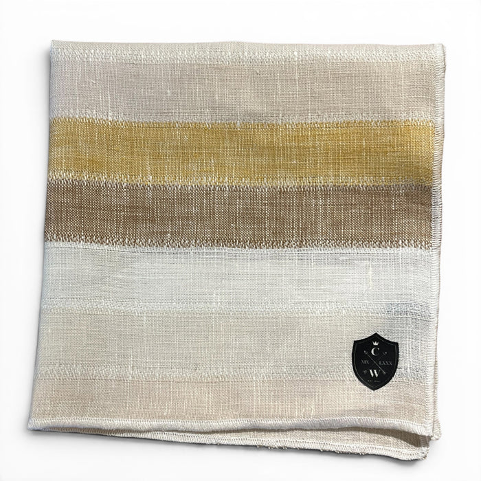 The CLIFTON WILSON - GOLD STRIPE LINEN POCKET SQUARE features beige, gold, brown, and white stripes with a black and white crest emblem at the bottom right corner. Made in the USA.