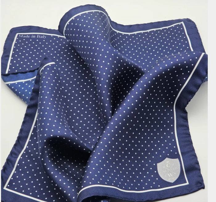 The CLIFTON WILSON - NAVY & WHITE POLKA DOT SILK POCKET SQUARE features a navy silk base with a white polka dot grid and thin white border, "Made in Italy" in the top left, and a small white shield emblem in the bottom right. Brand: CLIFTON WILSON.