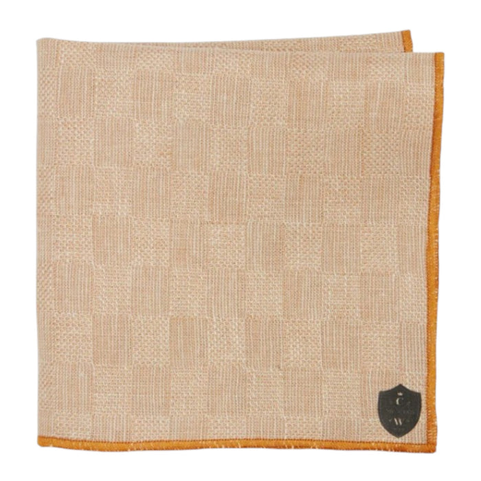 The CLIFTON WILSON - TAN CHECK LINEN POCKET SQUARE features a subtle check pattern, orange edging, and a small black shield emblem in one corner.