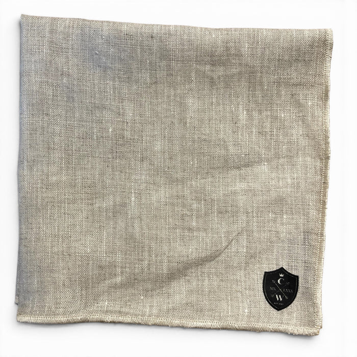The CLIFTON WILSON - TAN LINEN POCKET SQUARE features light tan textured linen with a black shield-shaped emblem of letters and symbols in one corner, reflecting the refined Clifton Wilson style.