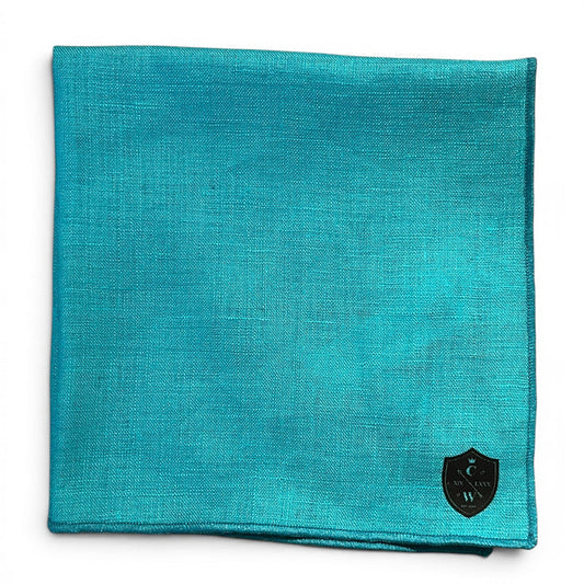 The CLIFTON WILSON - TEAL LINEN POCKET SQUARE features a black and silver shield logo in the bottom right corner. Made in the USA, it's shown neatly folded against a white background.
