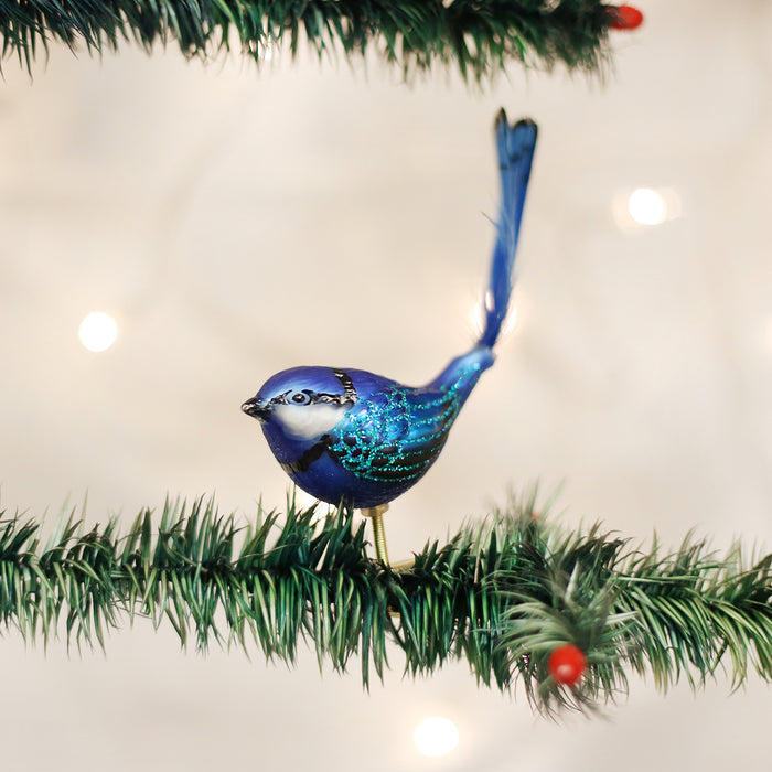 A blue and teal bird ornament with glittery wings perches on a green artificial pine branch decorated with small red berries. Warm white lights glow softly in the blurred background.