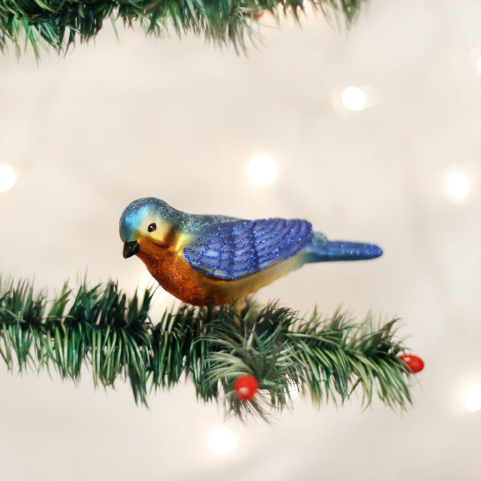A blue and orange bird ornament with glittery wings sits on a green Christmas tree branch decorated with small red berries, with soft white lights in the background.